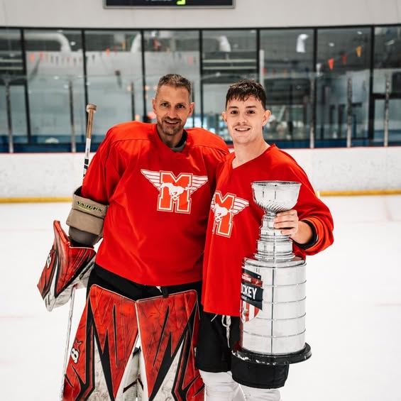 Two hockey players in red jerseys stand together on the ice, with one holding a silver trophy.
